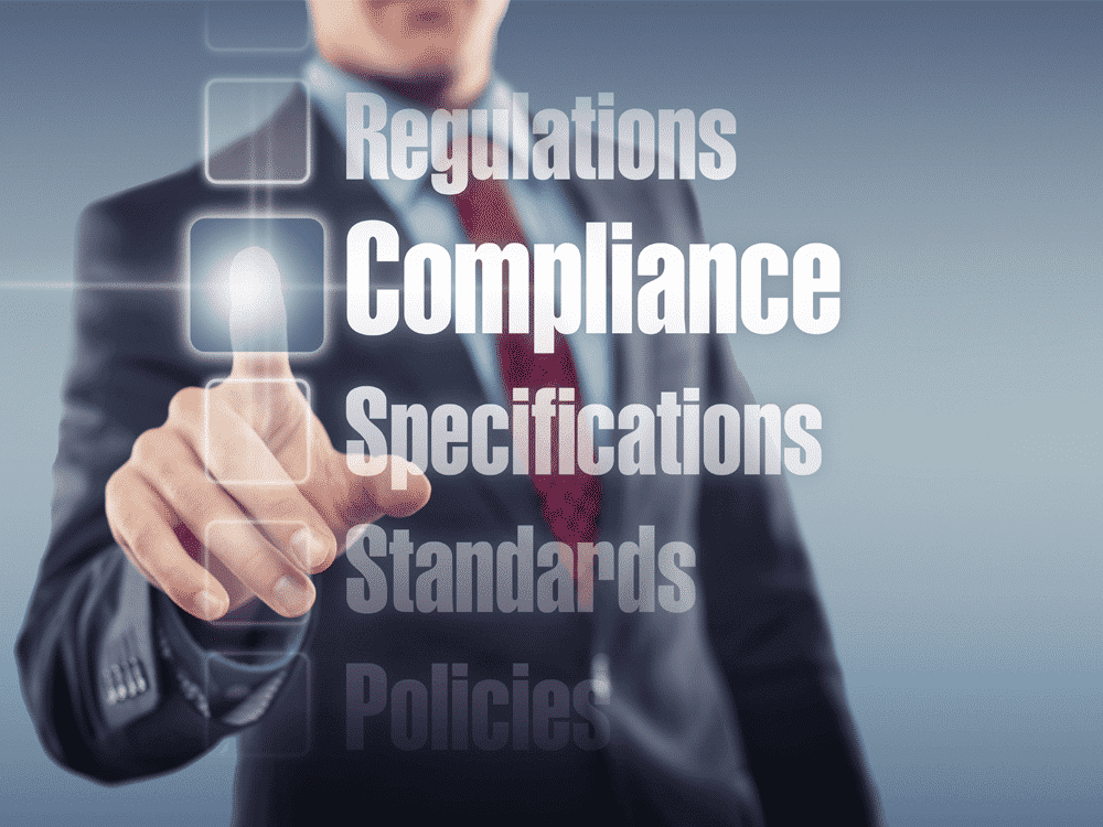 Man in black suit with red tie, pressing a button that says "Compliance" that is super imposed over his image