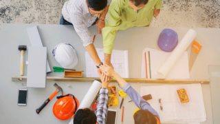 top view of group of engineer, technician and architect joining hands together with blueprint and construction tools on the conference table at construction site, teamwork, partner, industry concept