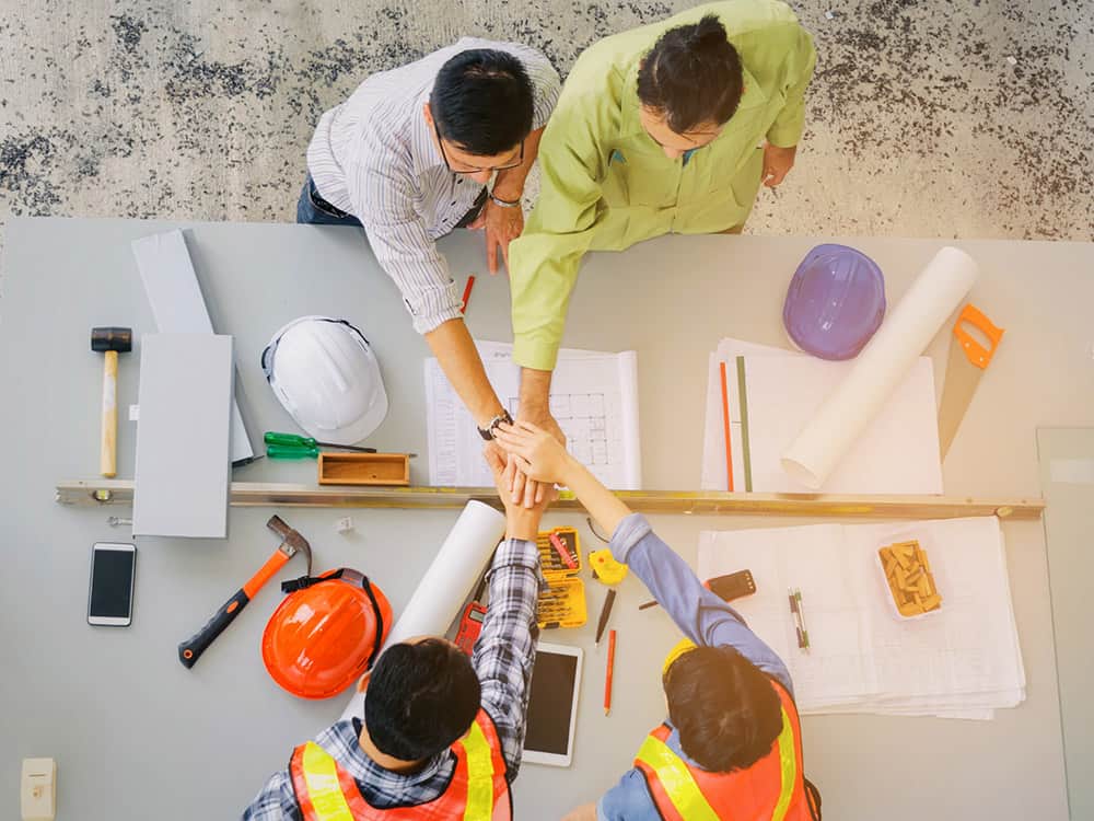 top view of group of engineer, technician and architect joining hands together with blueprint and construction tools on the conference table at construction site, teamwork, partner, industry concept