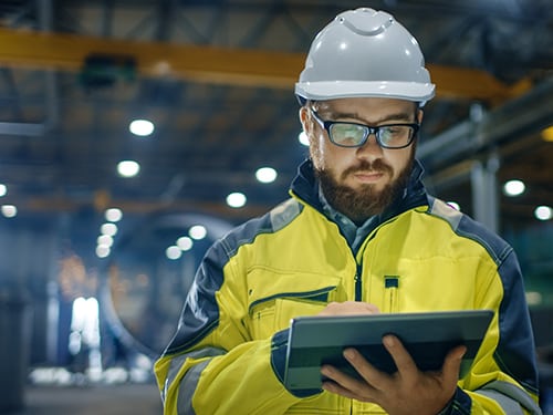 Industrial Engineer in Hard Hat Wearing Safety Jacket Uses Touchscreen Tablet Computer. He Works at the Heavy Industry Manufacturing Factory.